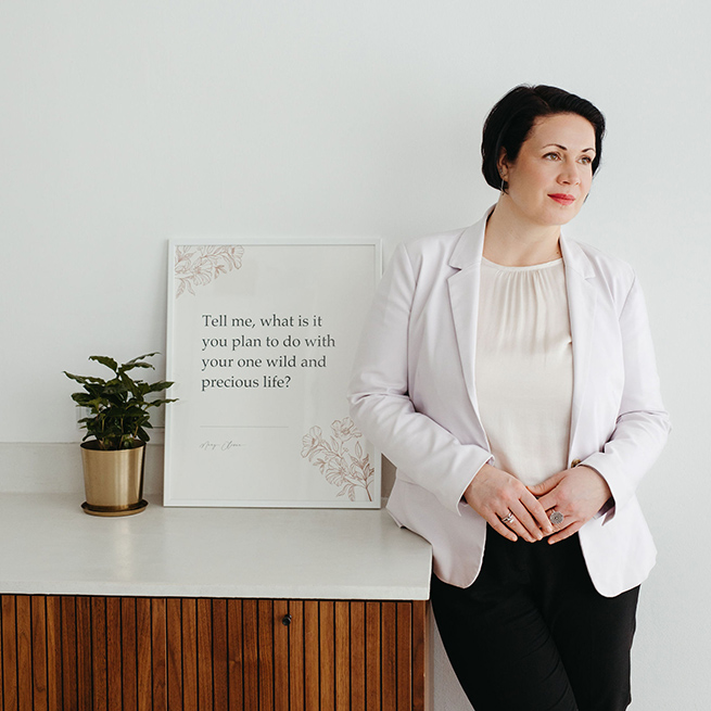 Coach Christine von Coaching Favorites steht angelehnt an einen Counter, darauf ein Bilderrahmen mit dem Zitat von Mary Oliver: Tell me, what is it you plan to do with your one wild and precious life?