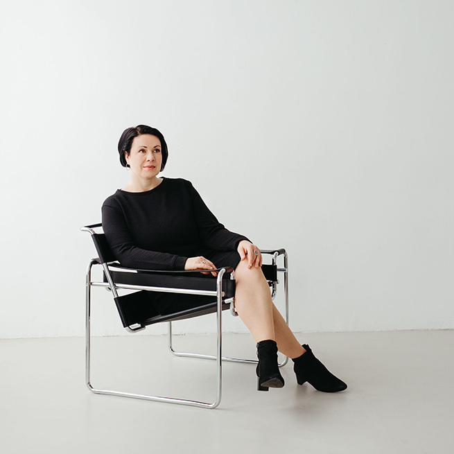 Coach Christine in einem eleganten schwarzen Kleid. Sie sitzt auf einem Stuhl und blickt nachdenklich in die Ferne.