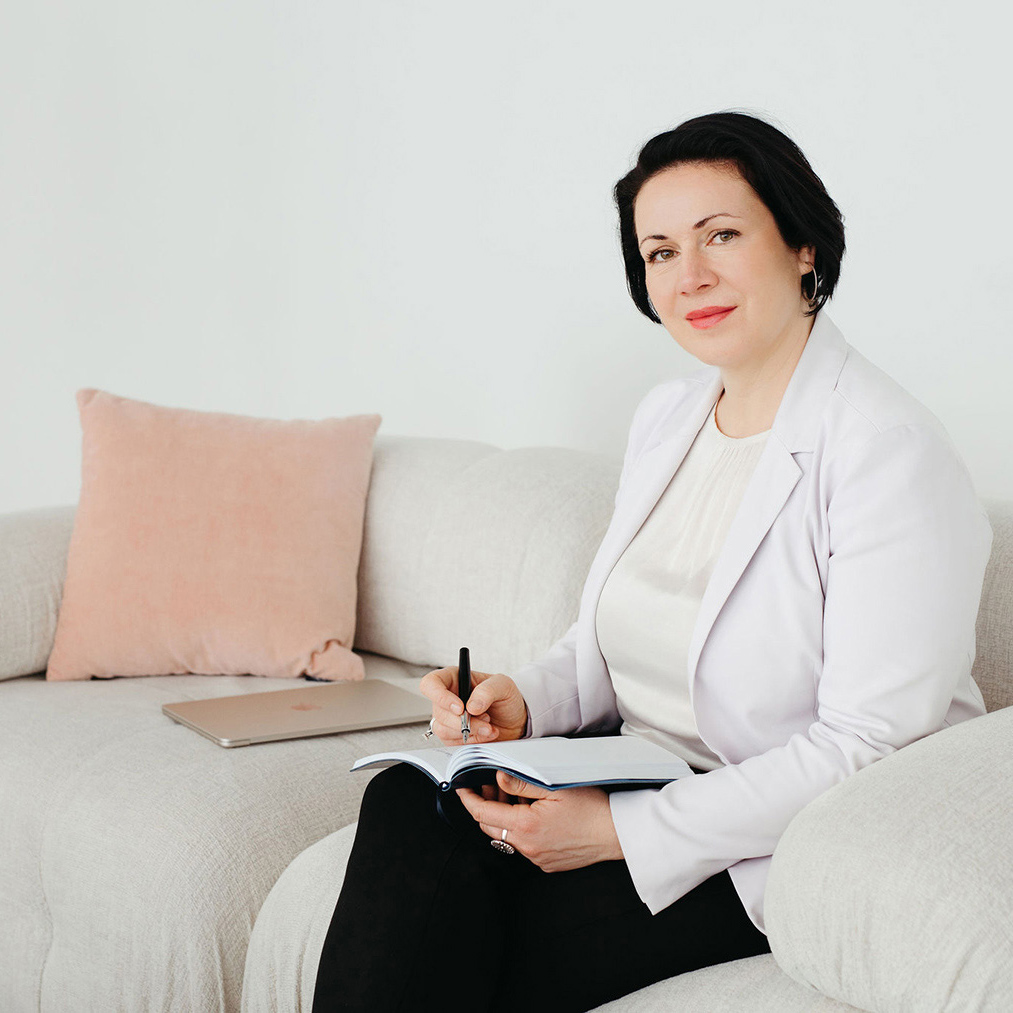 Christine bietet Coaching für Streberinnen an: Sie sitzt auf einem hellen Sofa, neben ihr ein Macbook und auf dem Schoß ein Notizbuch, in das sie mit Füller schreibt. Sie blickt freundlich in die Kamera.
