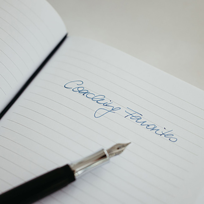 Ein Notizbuch mit dem Schriftzugn "Coaching Favorites" auf liniertem Papier, handgeschrieben mit blauer Tinte, daneben liegt ein eleganter Füller.