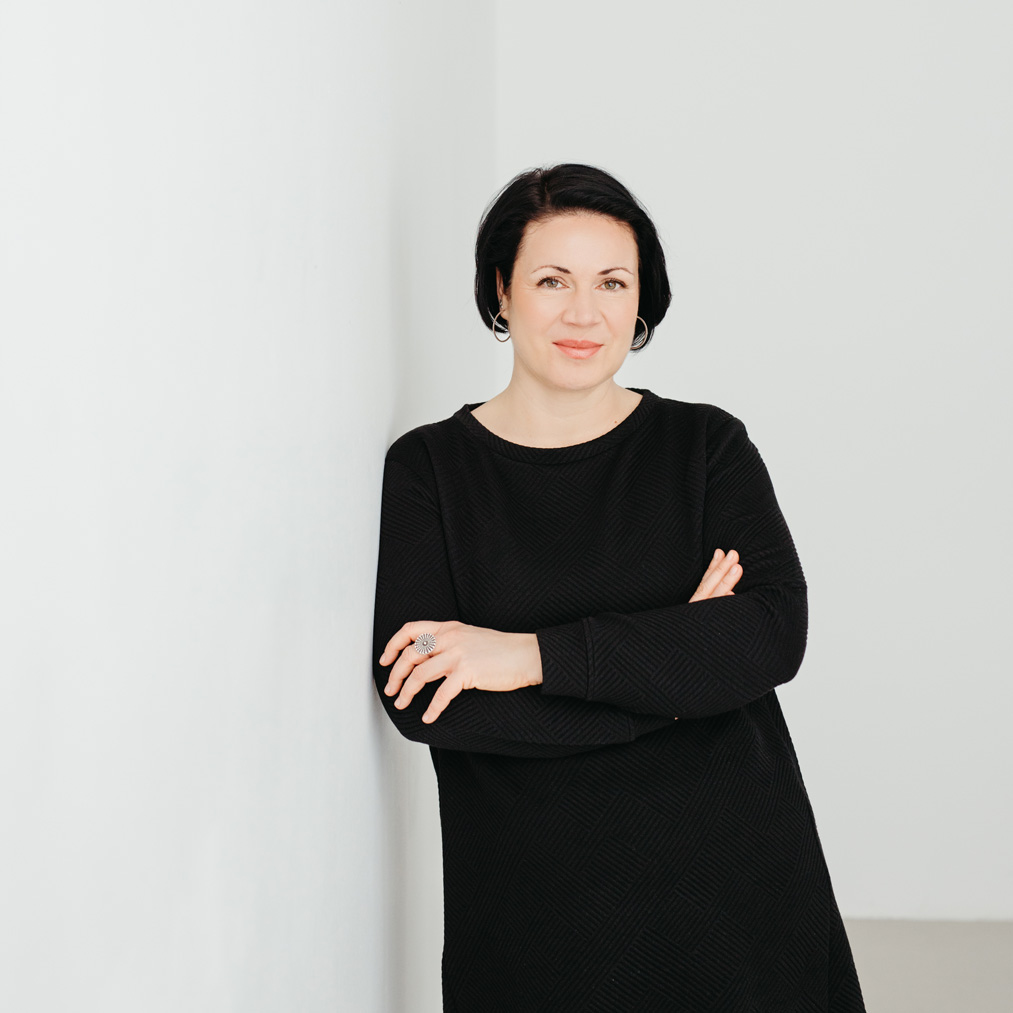 Christine bietet Coaching für Streberinnen an: Sie trägt ein elegantes schwarzes Kleid, steht an eine Wand gelehnt mit verschränkten Armen und lächelt.